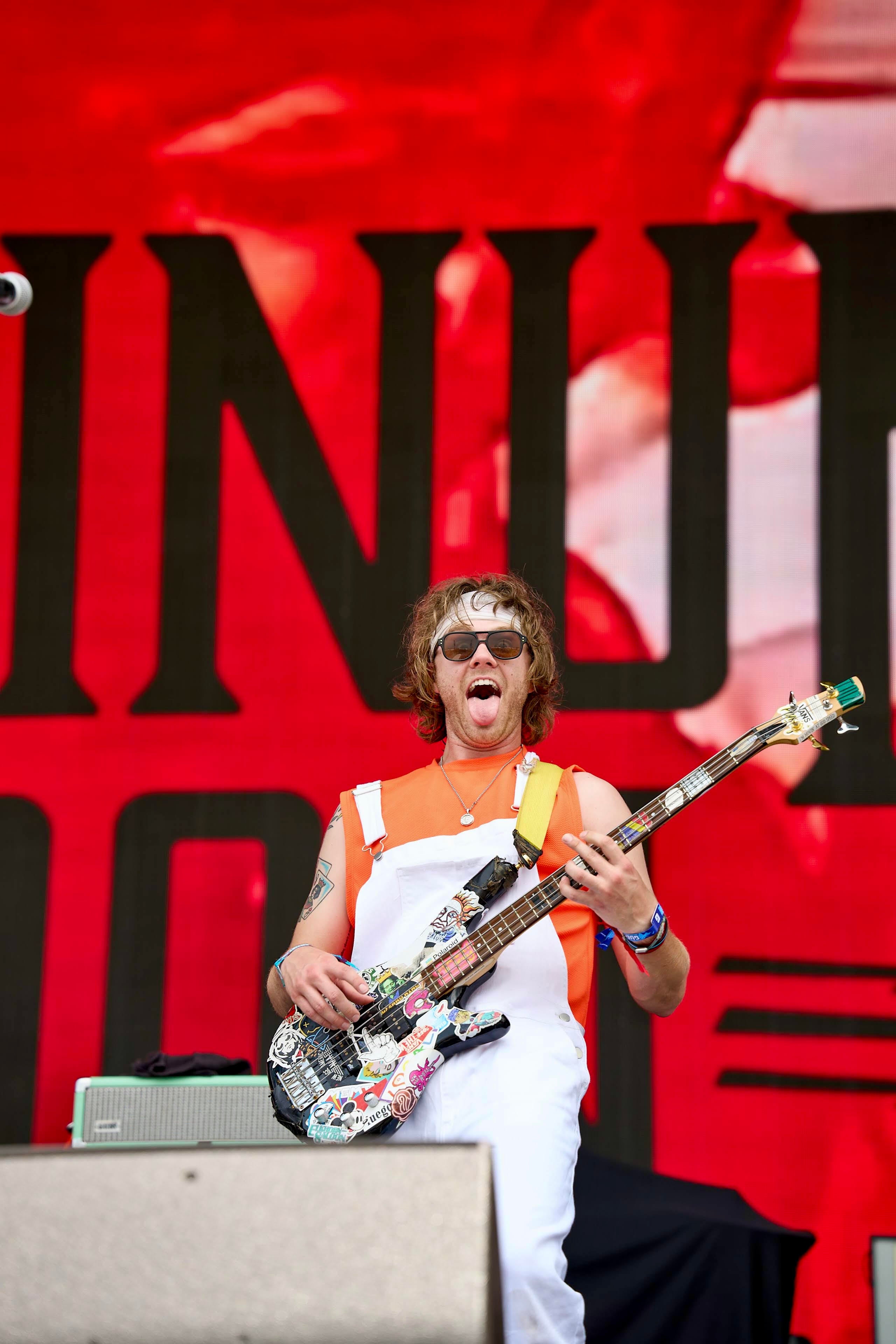 A person on a stage wearing white overalls and sunglasses, holding a colorful electric guitar in front of a large red and black backdrop.